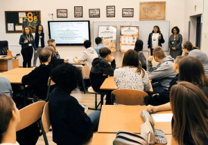 W sali lekcyjnej uczniowie siedzą w ławkach. Przed tablicą po lewej i prawej stronie stoją nauczyciele prowadzący zajęcia.