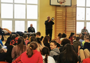 Na sali gimnastycznej na podłodze siedzą uczniowie. Z przodu przed nimi widać osobę grającą na puzonie oraz mężczyznę grającą na keyboardzie.