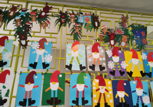 Na zdjęciu na kracie na korytarzu szkolnym wiszą prace uczniów. Są to mikołajki zdobione różnymi kolorami i różnymi ubiorami.
