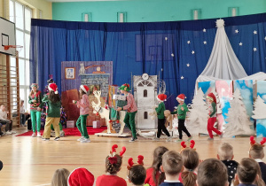 Na sali gimnastycznej odbywa się przedstawienie jasełek. Widać widownię, dekorację oraz biegnące elfy.