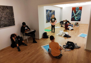 W pomieszczeniu galerii sztuki wśród obrazów na podłodze siedzą dzieci i malują swoje prace.