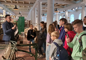 W sali muzealnej zgromadzone są dzieci przysłuchujące się wykładowi. Widać maszyny włókiennicze.