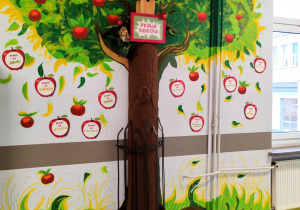 Na ściania na korytarzu szkolnym widać drzewo namalowane przez nauczyciela plastyki. Widać rownież jabłka. Na podłodze stoją kosze z jabłkami.