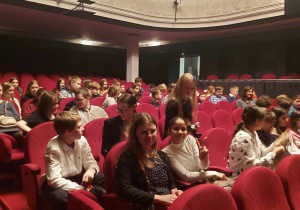 Na widowni w teatrze siedzą uczniowie klas szóstych. Wystrój wnętrza jest w kolorze czerwonym.
