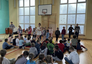 Na sali gimnastycznej na podłodze siedzą uczniowie. Przed nimi stoi osoba prowadząca zajęcia oraz dzieci uczące się kroków tanecznych.