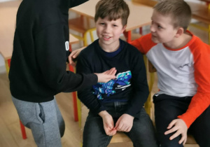 Scenka rodzajowa w klasie. Dwóch uczniów siedzi na krzesełkach. Trzeci uczeń stoi nad swoimi kolegami z założonym kapturem na głowie.