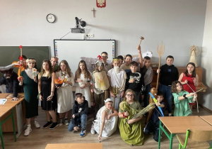 Zdjęcie grupowe uczniów przebranych za postacie z mitologii greckiej. Zdjęcie zrobione w sali lekcyjnej. Uczniowie stoją przed tablicą.