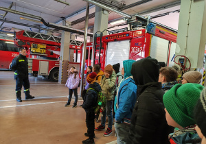 W garażu straży pożarnej stoją dzieci obok wozów strażackich. Widać strażaka prowadzącego zajęcia