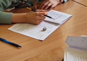 Na zdjęciu widać ręce dziecka rozwiązującego test. Na stole leży kartka z zadaniami, przybory piśmienne oraz karta odpowiedzi.