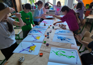Na stołach przykrytych papierem leżą prace uczniów w trakcie tworzenia. Widać dzieci dookoła stołów. W rękach trzymają sznurek umoczony w barwniku.
