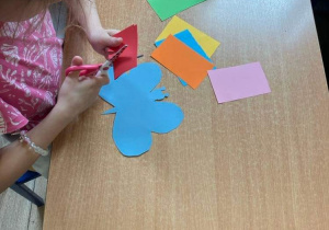 Na stole leżą karki papieru kolorowego. WIdać ręce dziecka wycinającego czerwoną kartkę. Na stole leży niebieski motyl wycięty z kartki.