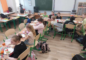 W sali lekcyjnej uczniowie siedzą w ławkach ustawionych w prostokąt i jedzą wspólnie śniadanie.