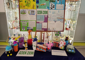 Na szkolnym korytarzu na stole stoją zwierzątka i stworki wykonane z surowców wtórnych. Z tyłu znajduje się tablica z plakatami zawierającymi różne informacje ekologiczne.