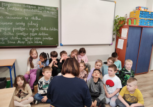 W sali szkolnej uczniowie siedzą pod tablicą i słuchają nauczyciela czytającego książkę.