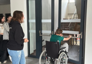 Na korytarzu stoją uczniowie. Przyglądają się koledze siedzącemu na wózku inwalidzkim i próbującemu otworzyć drzwi.