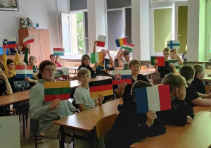 W sali szkolnej siedzą uczniowie, w rękach nad głowami trzymają flagi krajów europejskich