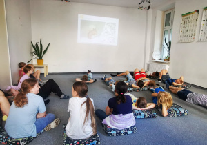 W pomieszczeniu dzieci leżą na podłodze, oglądają prezentację o zwierzętach. Widać również osobę prowadzącą zajęcia