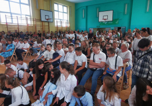 Na sali gimnastycznej uczniowie i rodzice siedzą na krzesełkach.