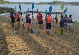 Na piaszczystej plaży w okręgu stoją uczniowie, trzymają wiosła. Są ubrani w kapoki. W dali widać jezioro.