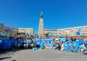 Na placu Wolności grupa młodzieży wraz z opiekunami stoi pod pomnikiem Tadeusza Kościuszki. Wszyscy trzymają niebieskie balony, niektórzy są ubrani w niebieskie koszulki.