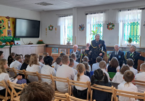 Na świetlicy szkolnej na krzesełkach siedzą uczniowie ubrani w stroje galowe. Widać kombatantów uczestniczących w spotkaniu ubranych w mundury oraz panią dyrektor.