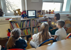 W bibliotece szkolnej dzieci siedzą na krzesełkach. Widać nauczycielkę opowiadającą o misiach. Widać również laptop z wyświetloną prezentacją o misiach. Na parapecie okiennym stoją misie.