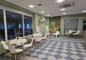 Na zdjęciu widać pomieszczenie pomalowane na oliwkowy kolor, na jednej ze ścian znajduje się fototapeta, na której widać leśną ścieżkę,stoją cztery białe stoły i szesnaście krzeseł