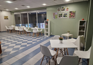 Na zdjęciu widać pomieszczenie pomalowane na oliwkowy kolor,stoją cztery białe stoły i szesnaście krzeseł, na ścianie wiszą zdjęcia przyrodnicze, stoją również dwa regały z książkami